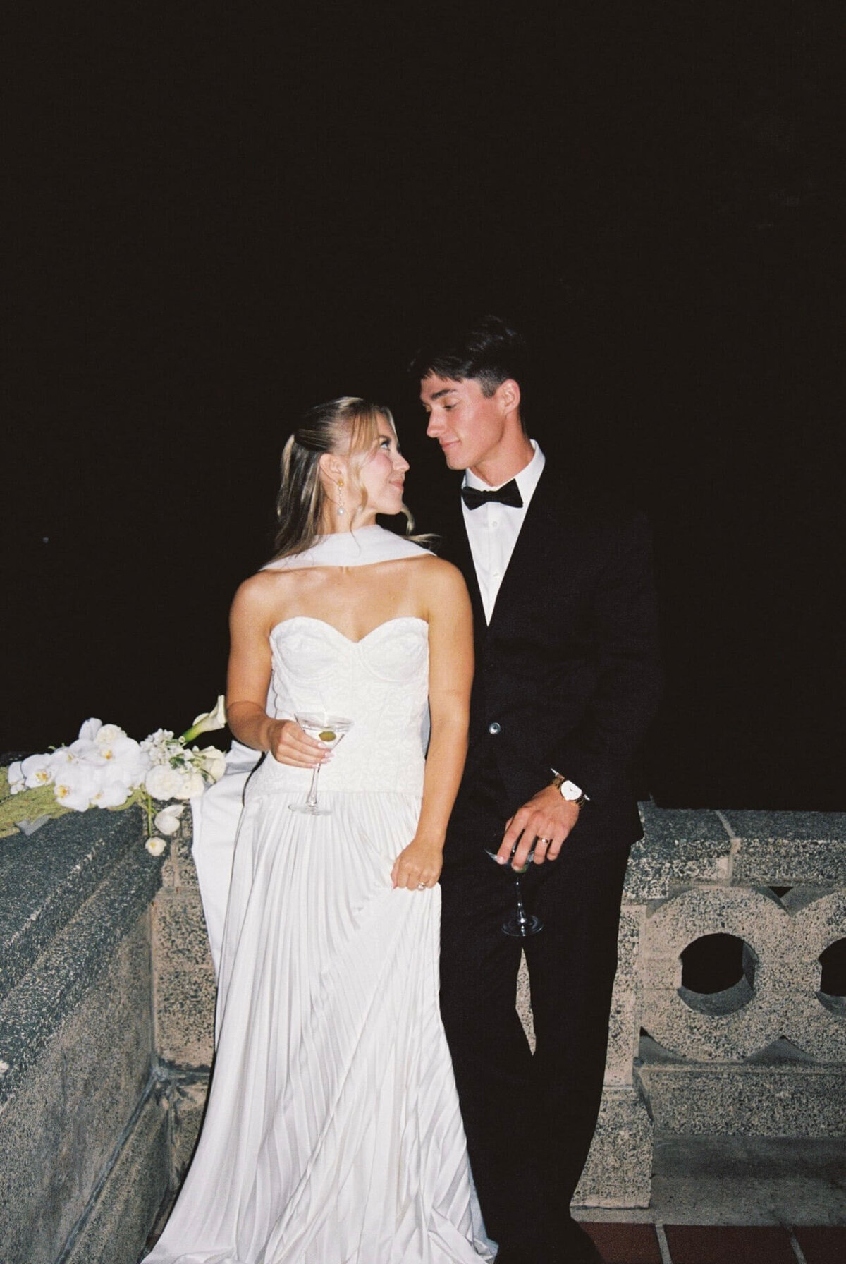 Newlyweds stand against a stone railing leaning on each other and holding martinis