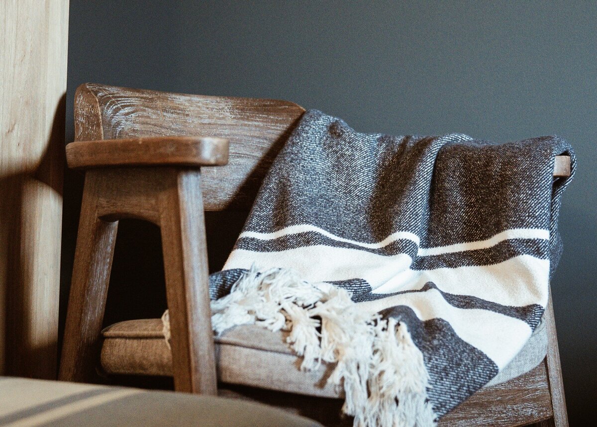Wooden barrel chair with cushion and woven blanket next to denim painted ottoman against dark blue green painted wall