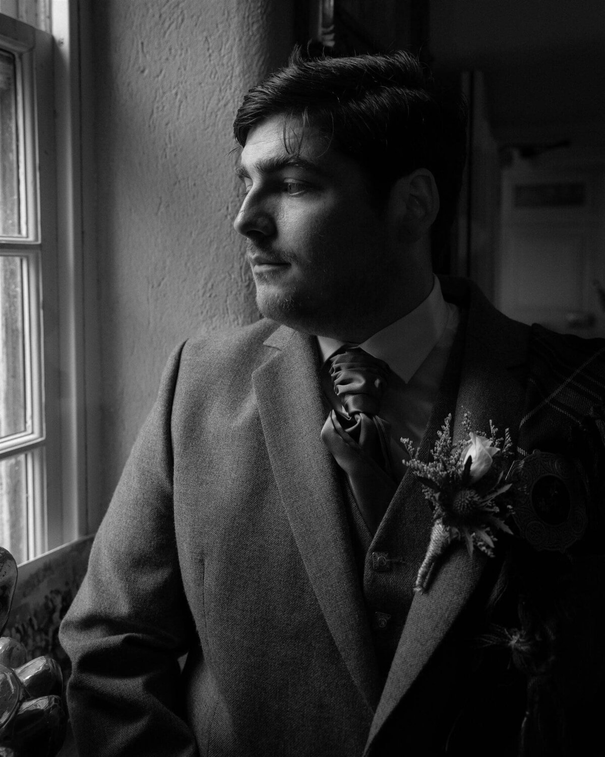 Groom looks out of a window. he wears a thistle on his jacket.
