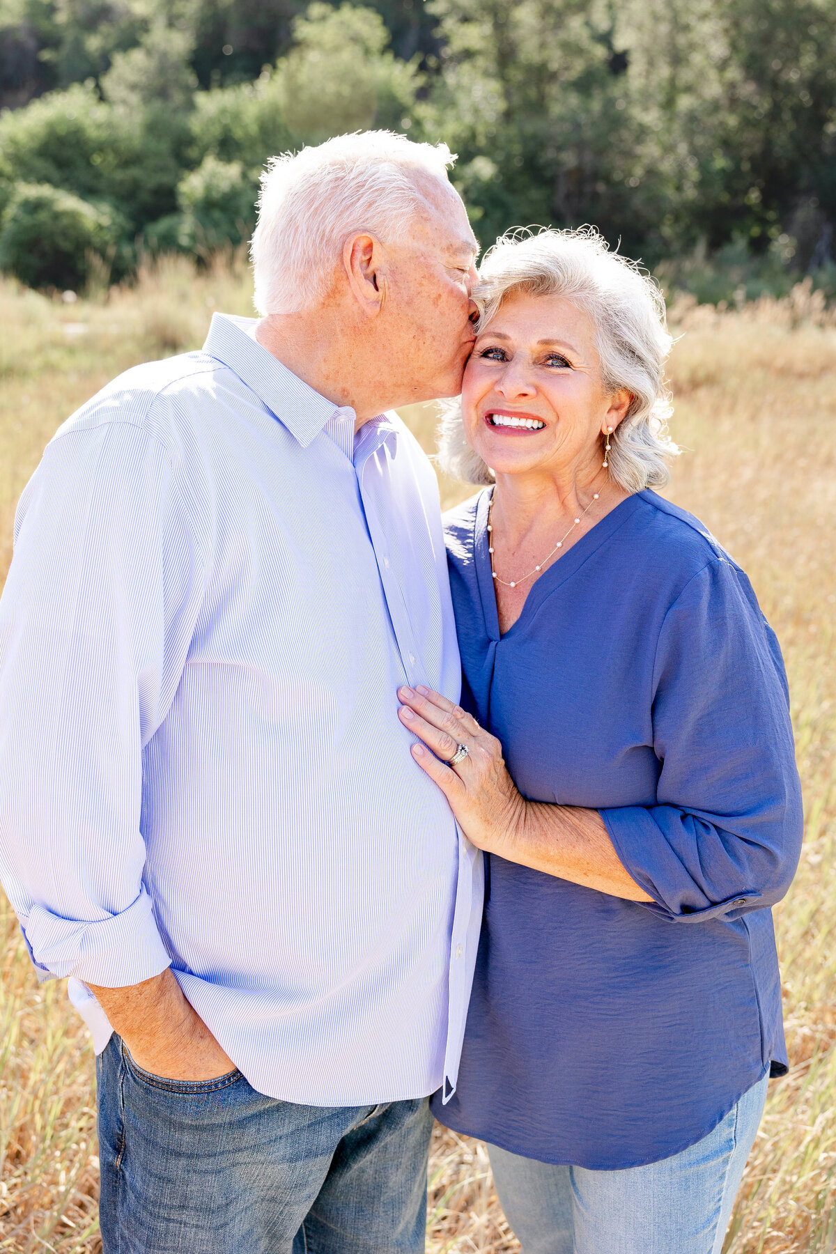 An elderly man giving his wife a kiss on the forehead as she smiles, both standing in a scenic outdoor setting.