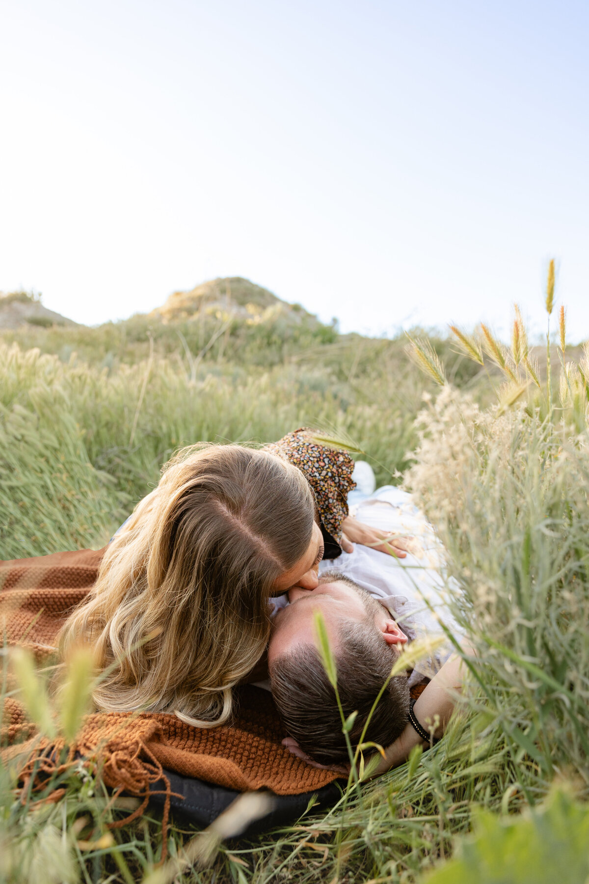 A woman kisses her fiancé while lying together on a blanket in a lush, grassy field. The soft natural tones of the setting create a warm and intimate atmosphere.