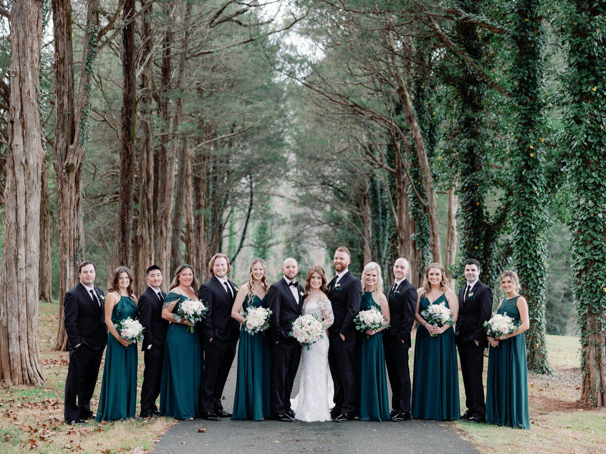 A bridal party of bridesmaids , groomsmen, and the couple stand together in a line underneath tall trees covered in ivy