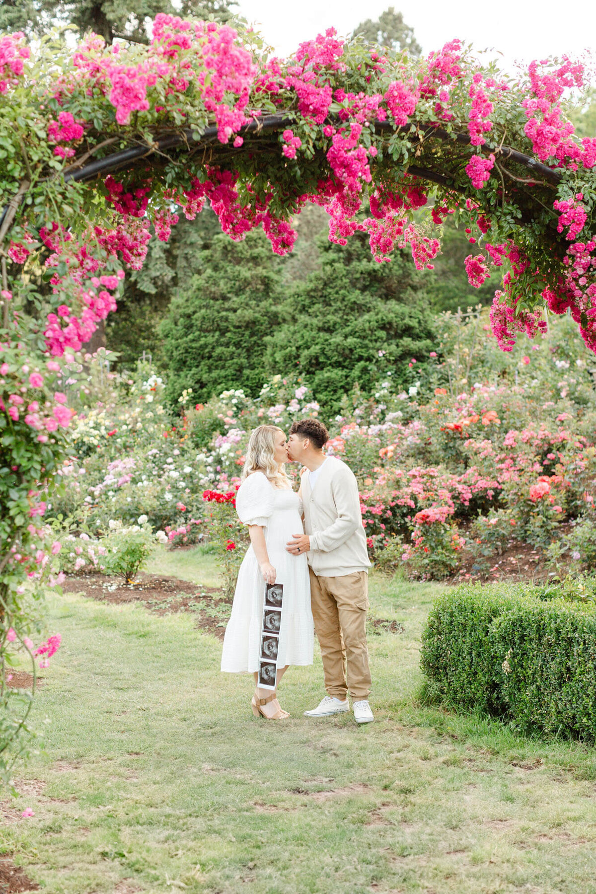 Pregnant Woman and Man share a kiss while woman holds ultrasound photos down by her side at the Portland Rose Gardens.