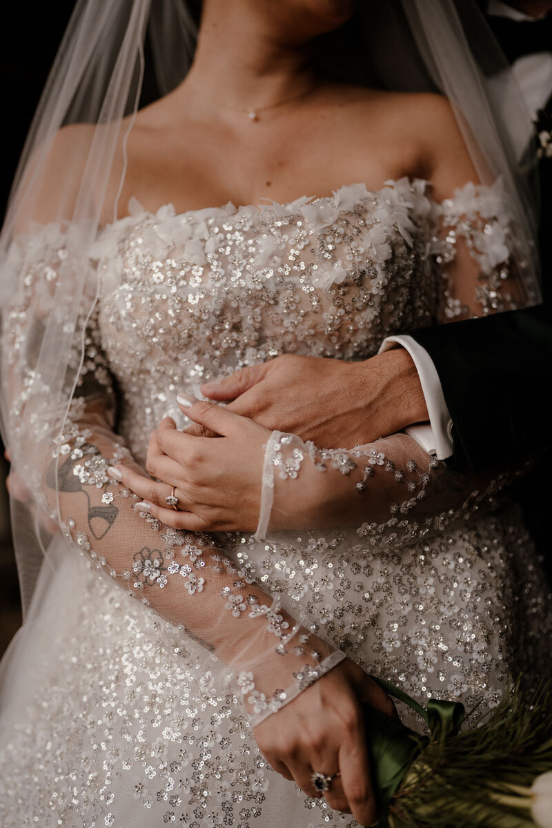 Détail des mains d’un couple de mariés enlacés, robe brodée de sequins, photo capturée lors d’un reportage photo et vidéo en Haute-Savoie.