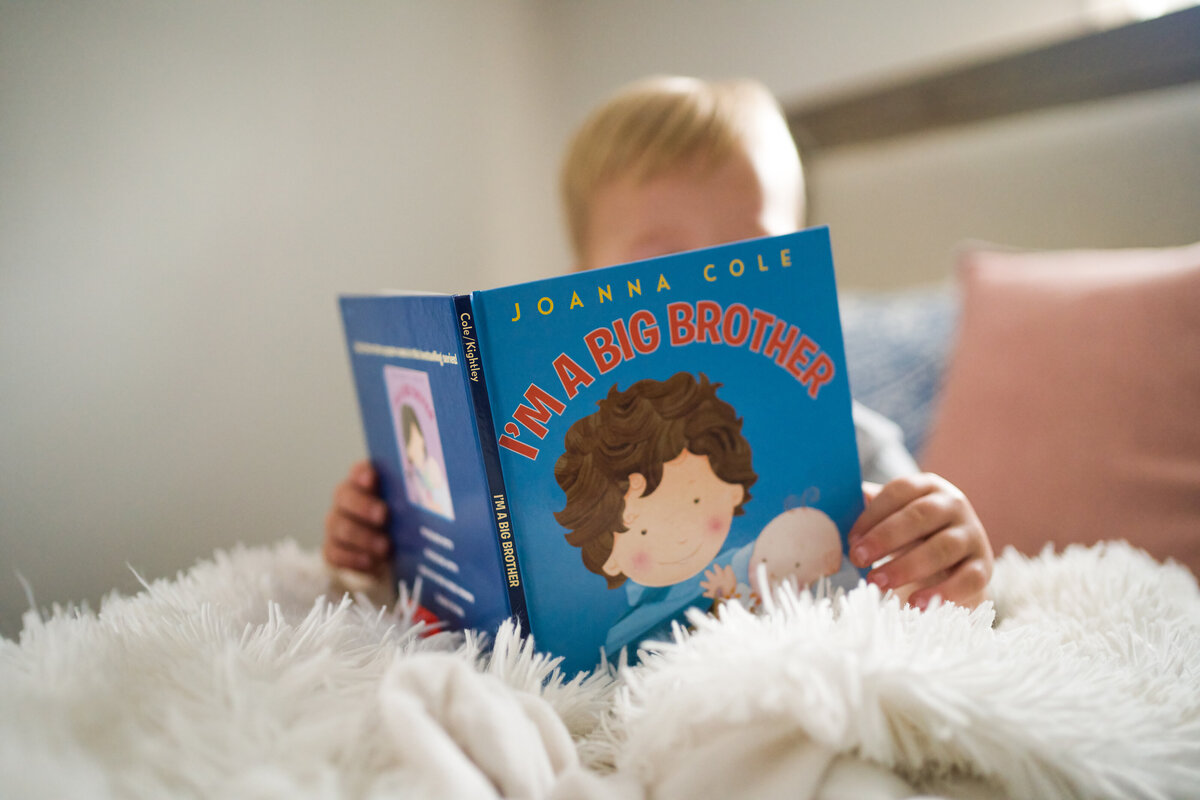 Toddler boy reading book about being a big brother during newborn picture session for his new little sister