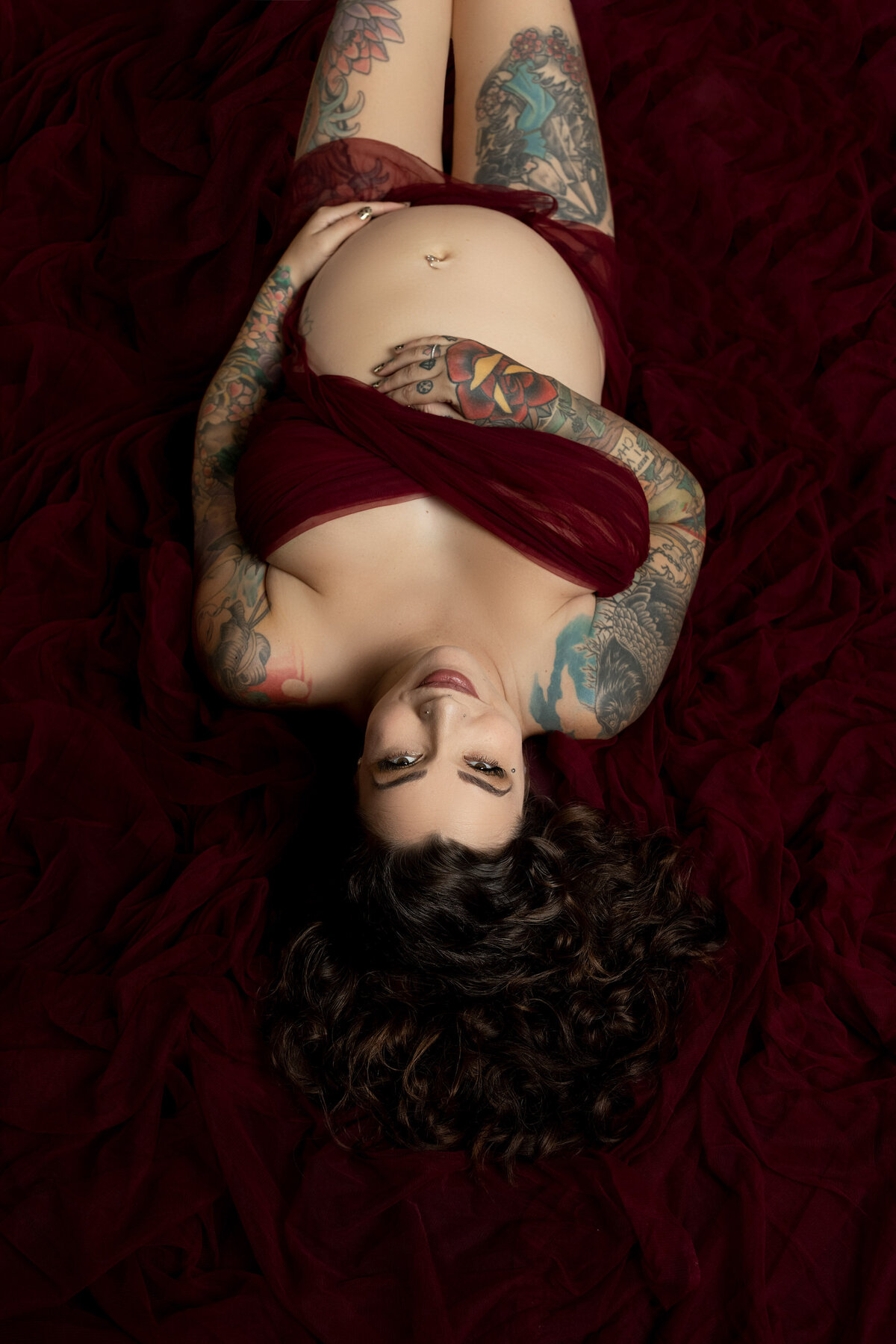 Expectant, tattooed mom in London, Ontario maternity studio photo session. Mom laying on burgundy velvet with only breasts covered. Colourful tattoos are prominent.