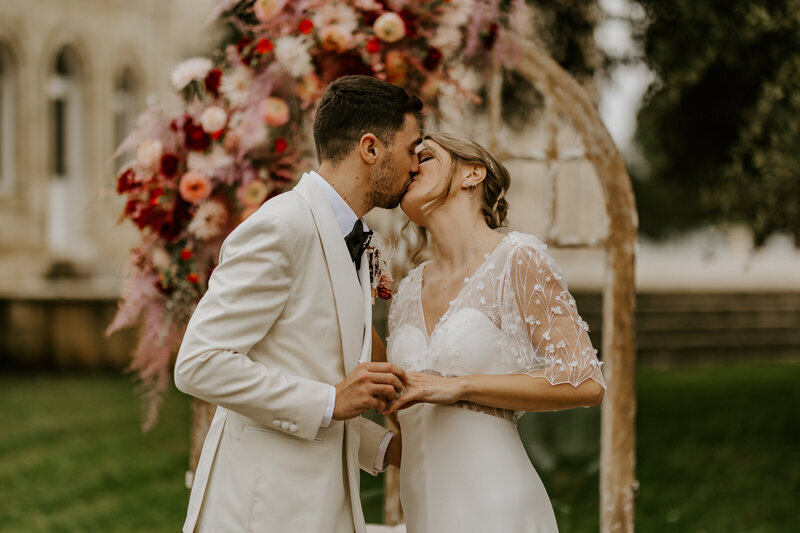 Mariés se faisant un bisou et passant en même temps la bague au doigt. Décor nature et arche en bois surmontée d'une composition florale. Shooting pour le workshop de photographie de mariage organisé par Laura.