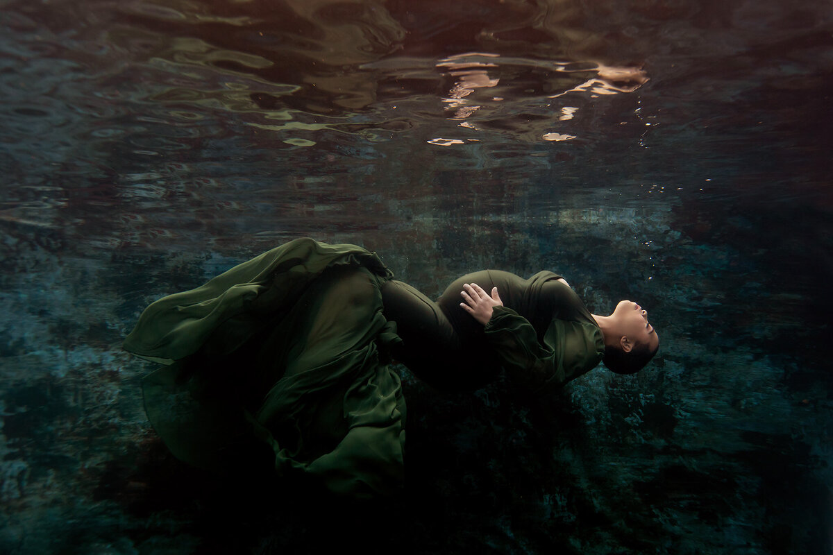 Underwater maternity photo featuring a woman elegantly arching backward in a flowing dark green gown, her body gently curving against a textured blue-green backdrop. The peaceful underwater environment highlights the artistry of this Dallas underwater maternity photographer, capturing the grace and tranquility of the moment.