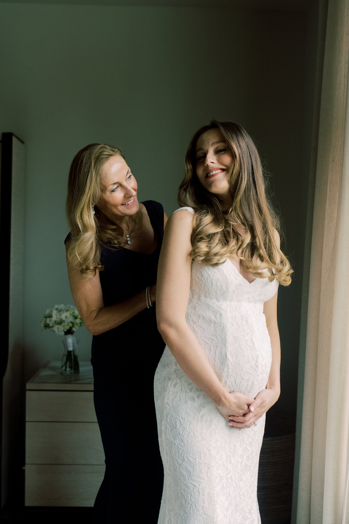 A candid, color photograph of a mother lovingly helping her pregnant daughter prepare for her bridal day, capturing the joy and tenderness of their bond.