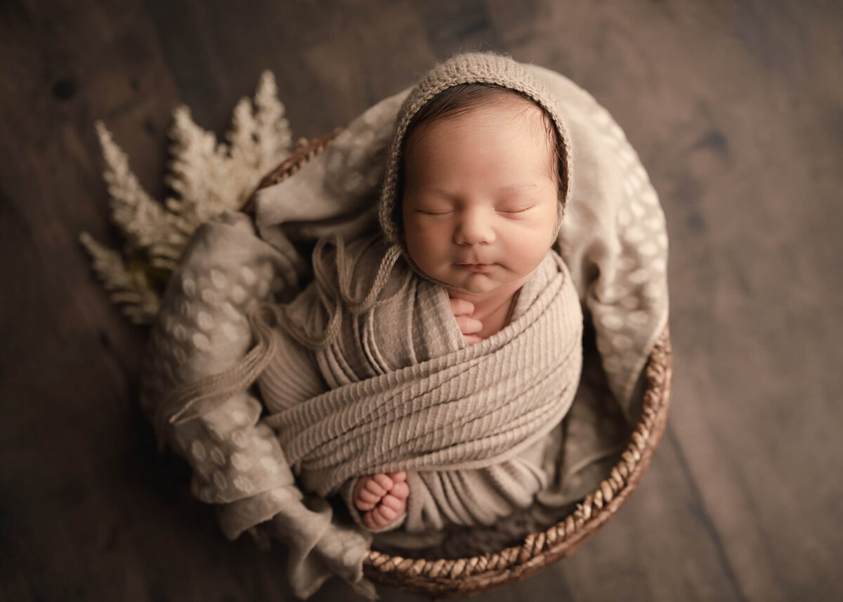 Aerial image. Baby swaddled and placed in a basket for a newborn photoshoot. Baby's fingers and toes are peeking out of the wrap. Baby is wearing a matching bonnet and sleeping peacefully. Captured by Lake Elsinore newborn photographer Bonny Lynn Photography.