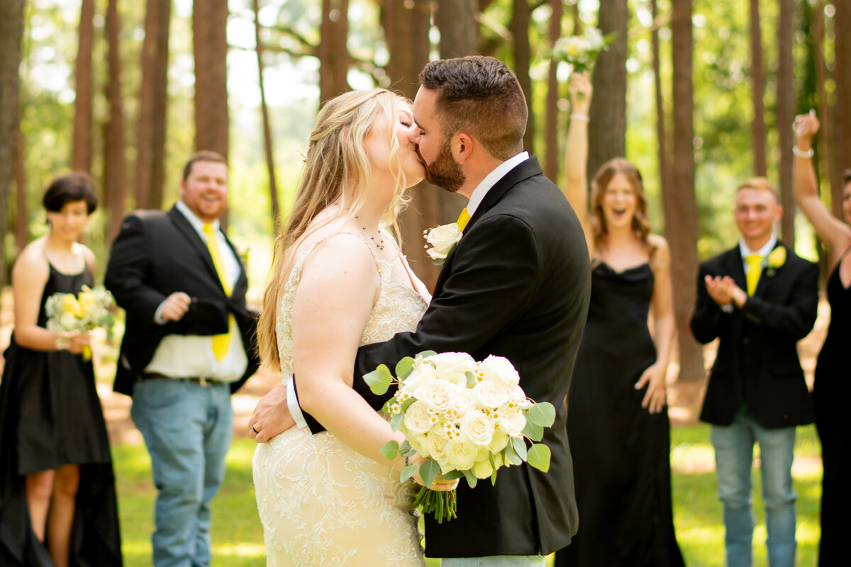 Witness the heartfelt moment as the bride and groom share a passionate kiss with their wedding party behind them at The Springs Event Venue in Magnolia, Texas, a celebration of love and unity