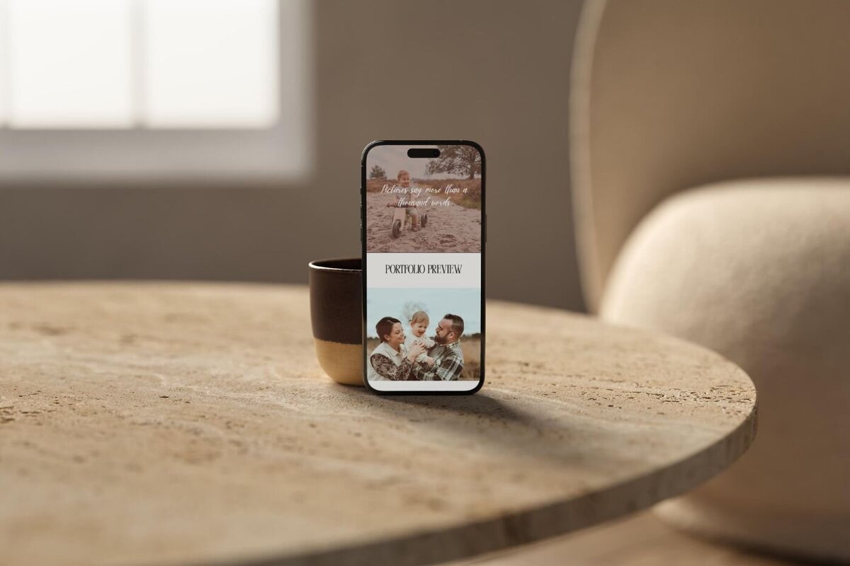 Smartphone standing on a brown-and-gold ceramic cup placed on a round stone coffee table, screen showing a photography portfolio preview with a smiling family in a sunlit field.