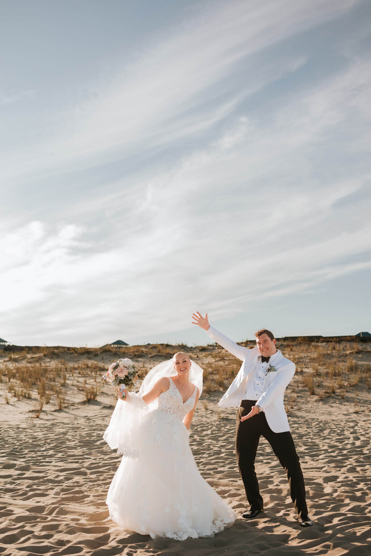 A joyful bride and groom pose playfully on a sunny beach, the bride holding a bouquet and the groom gesturing animatedly. A LBI wedding.