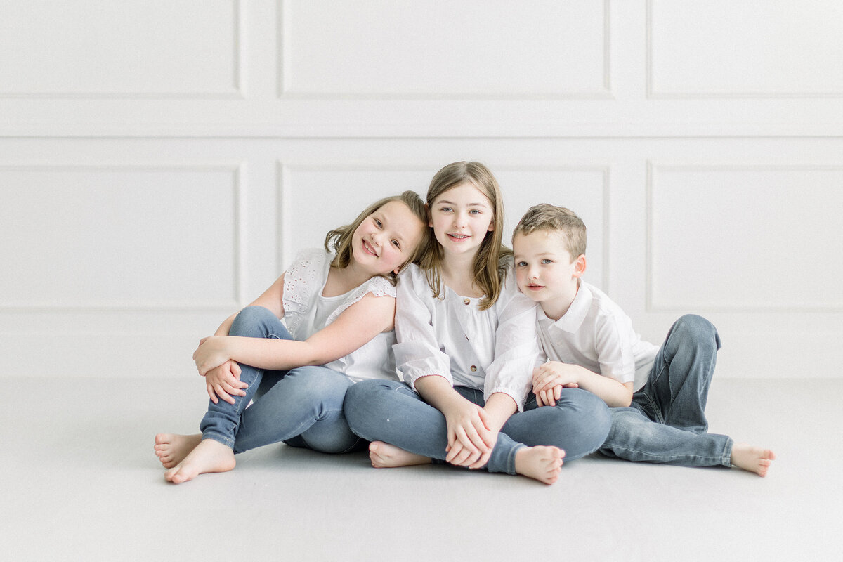 A photo of 3 young siblings sitting on the floor leaning on each other at a photography studio in Fort Worth TX.