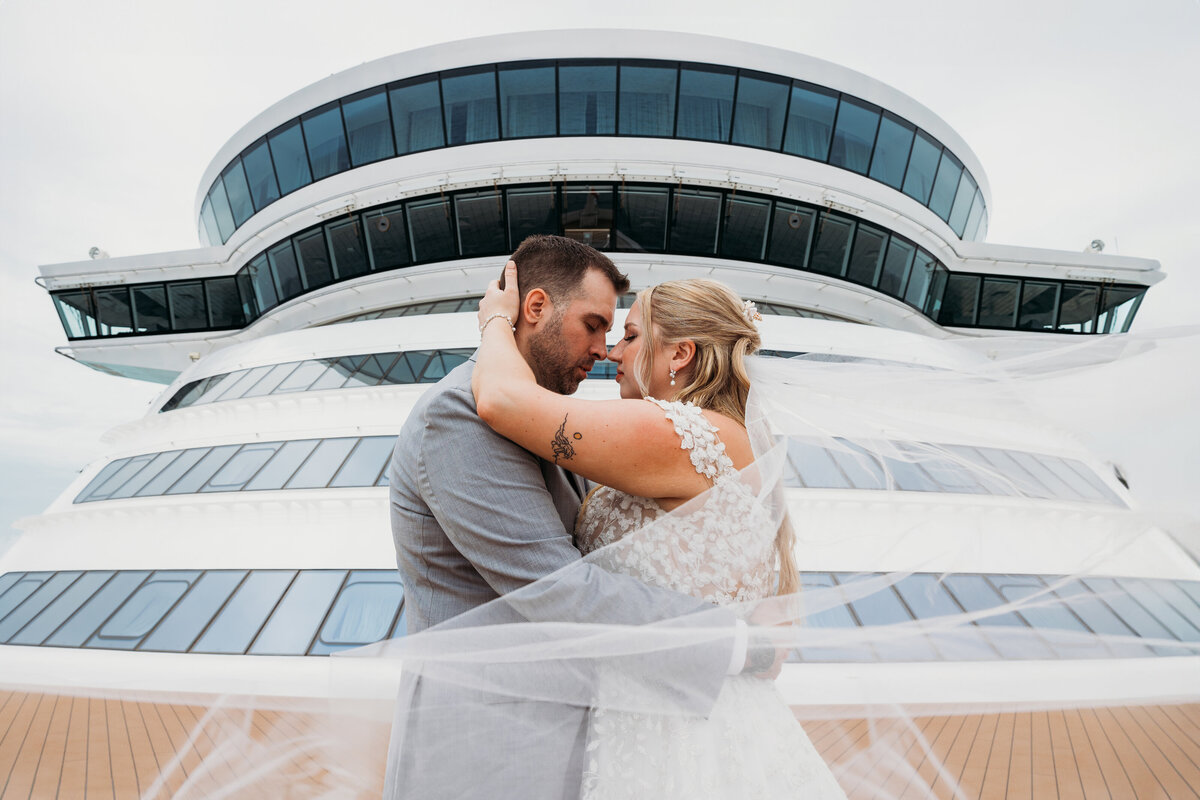 The couple embraces on the outer deck of the Disney Wish, with the bride’s veil floating in the breeze during their wedding day.