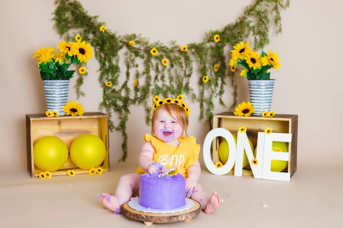Cake Smash Photographer, a baby girl smiles before cake and a backdrop full with sunflowers