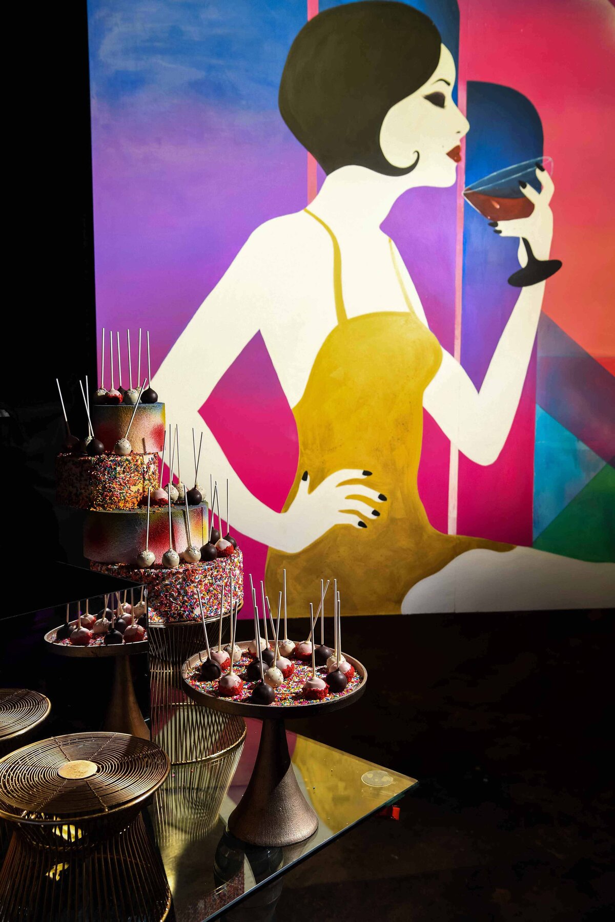 Cake and cake pops are displayed on a table with a painting in background. The image is lit by professional lighting for a creative food product shoot