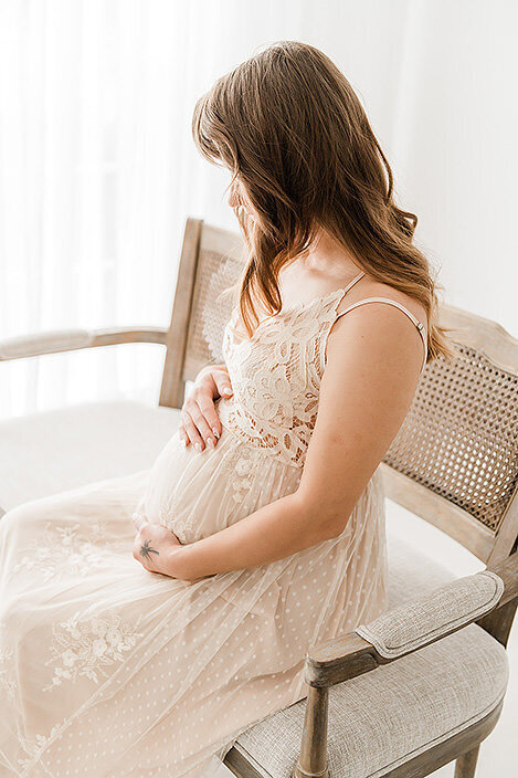 Natural and graceful portrait of an expecting mother seated in a vintage-style wooden chair near soft window light.
