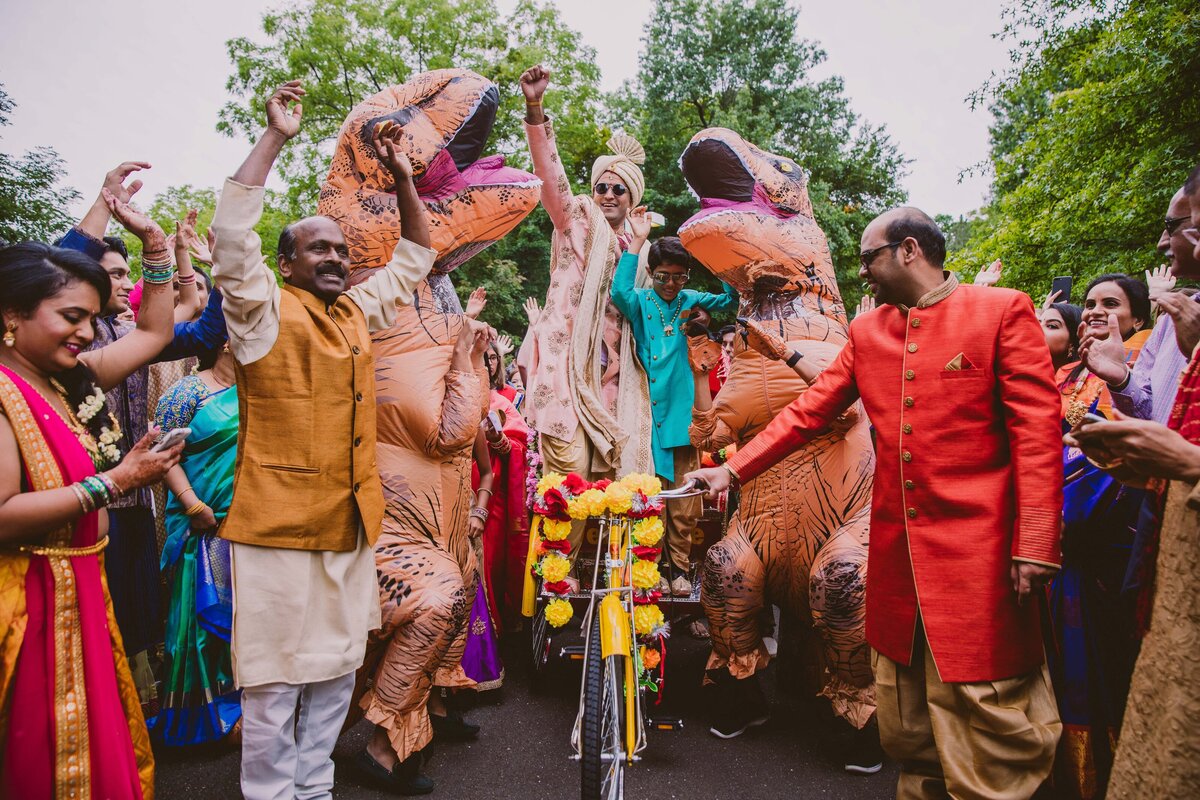 An indian groom dancing at his baraat on a decorated bike with his family and inflatable dinosaurs