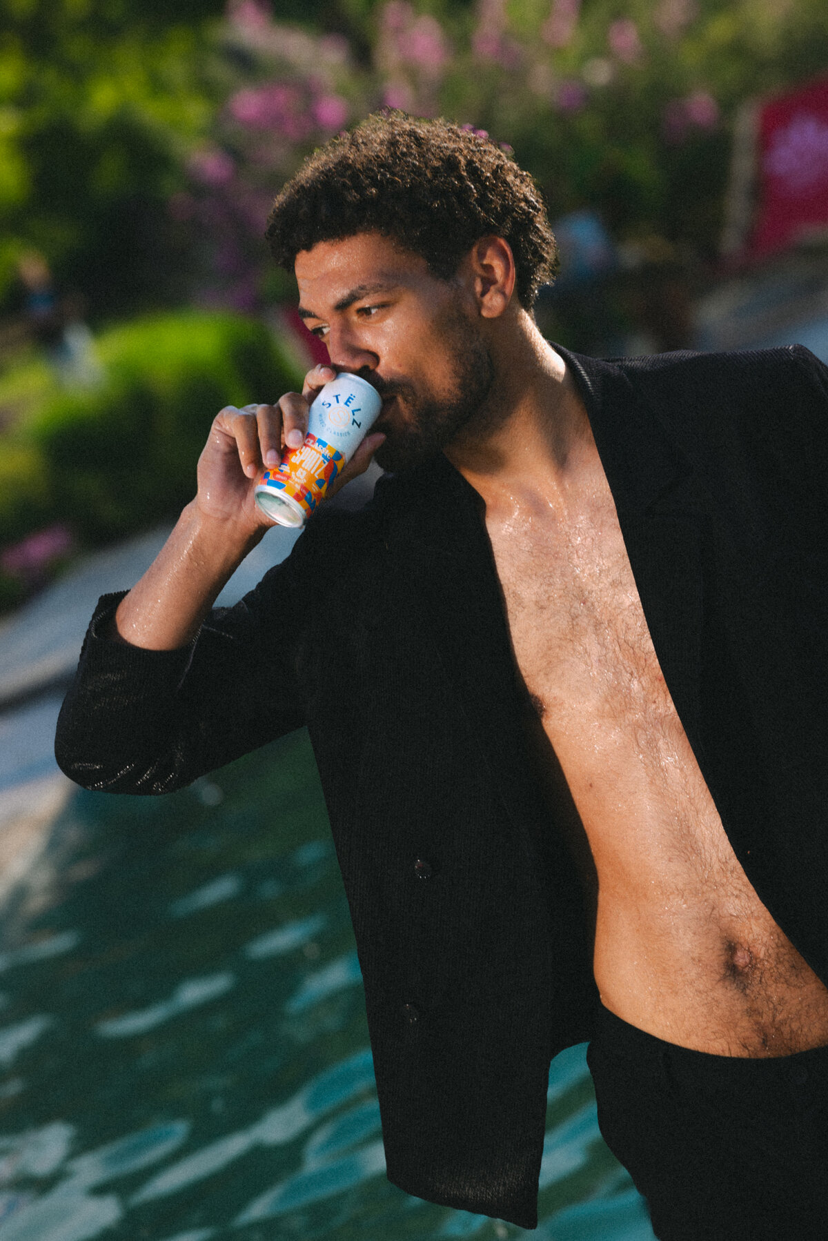A man with wet hair and a stylish black jacket drinks from a STËLZ Classic Spritz can, standing by a pool. The image exudes a vibe of sophistication and leisure, perfectly blending fashion with a refreshing drink. Photography by Bo Eppink.