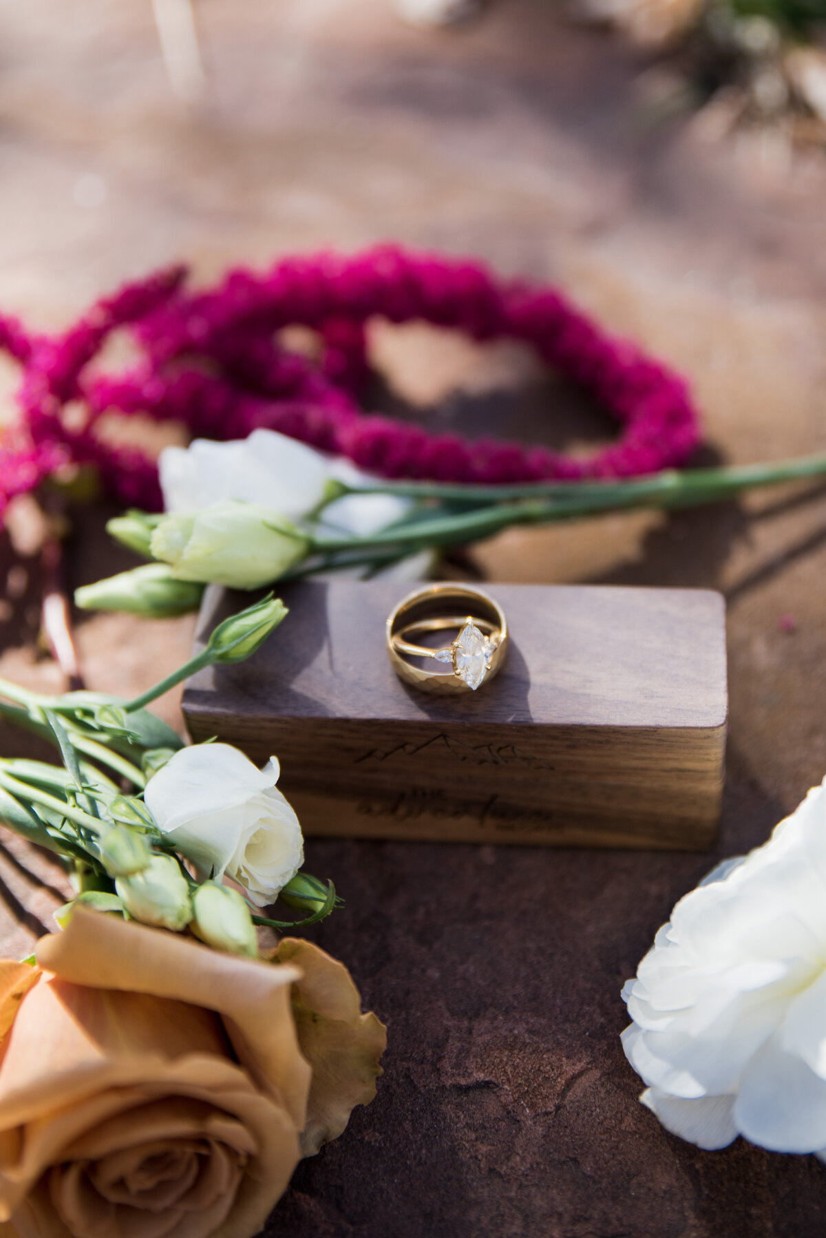 A diamond engagement ring and men's wedding band sit on a wooden box with an orange rose in the foreground.