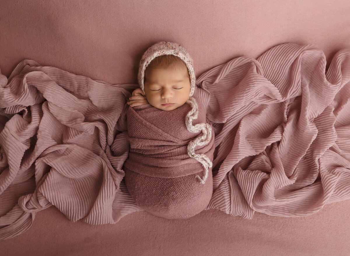 Brooklyn newborn photoshoot. Aerial image. Newborn baby girl is wrapped in a dusty rose knit swaddle with her hands peeking out and resting on her cheek. She is wearing a blush knit bonnet and laying atop of dusty rose fabric. Captured by premier Brooklyn NY newborn photographer Chaya Bornstein Photography.