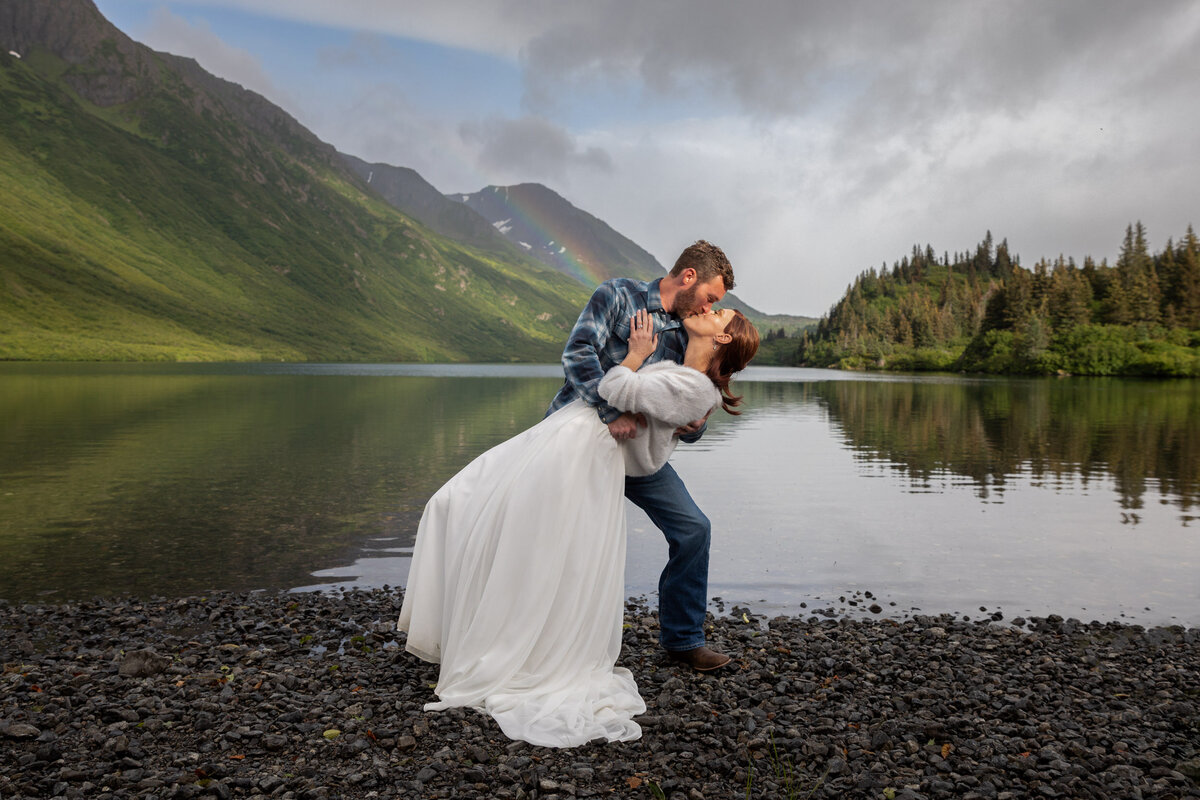 A groom dips his bride and kisses her in front of a lake in Alaska