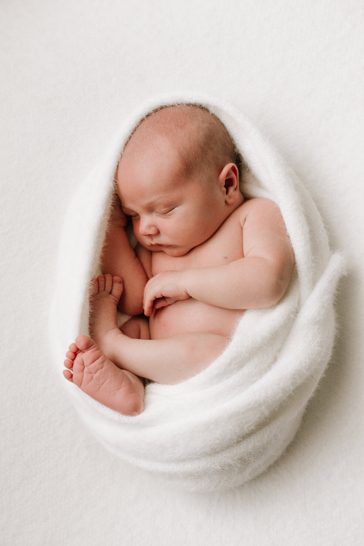 A newborn baby peacefully sleeping, swaddled in a soft white wrap with legs curled in a natural pose, resting on a textured white blanket. The soft artificial light used in the Sydney studio highlights the baby's delicate skin and tiny features.