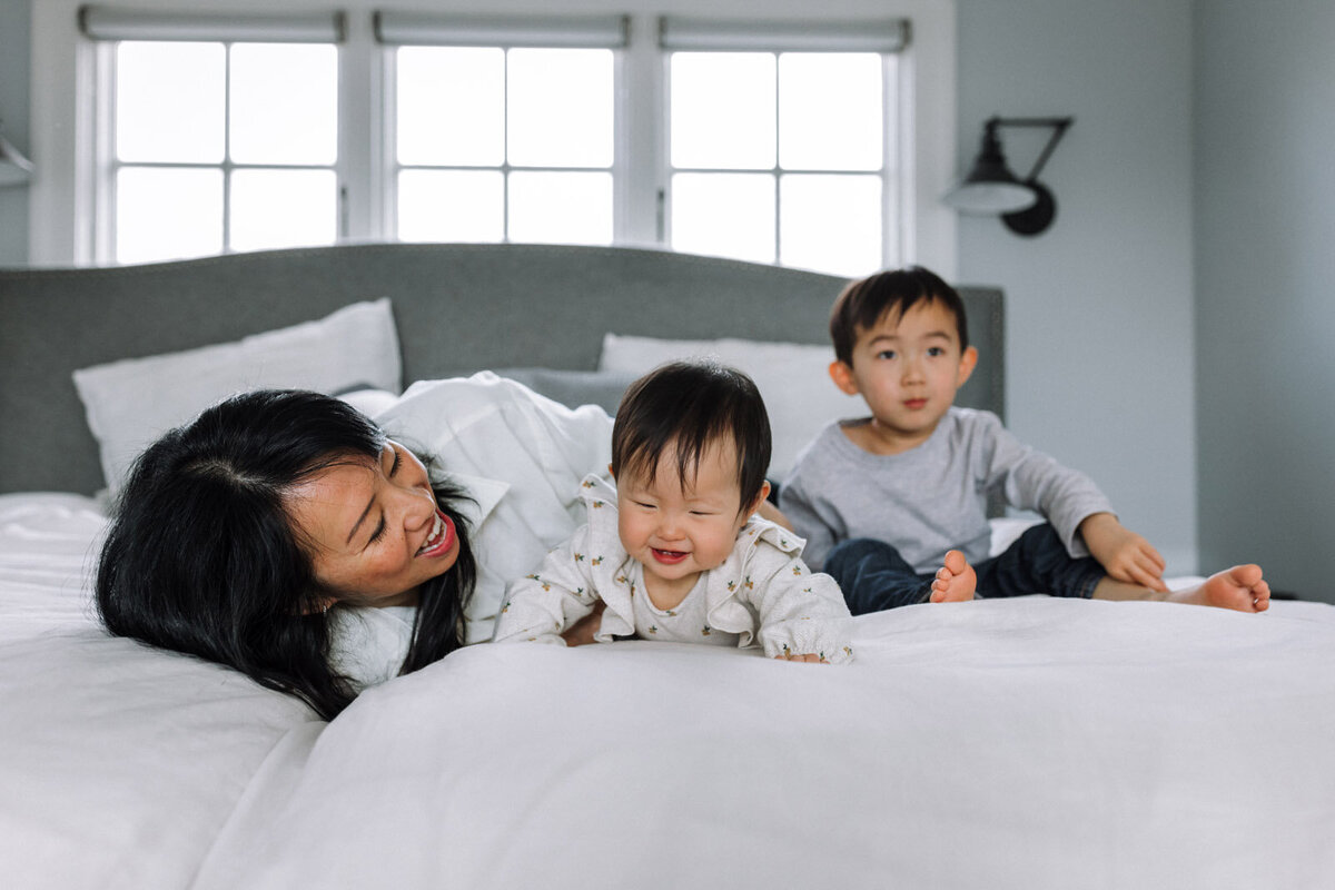 mom with toddler and baby on bed playing and interacting as son sits in the distance