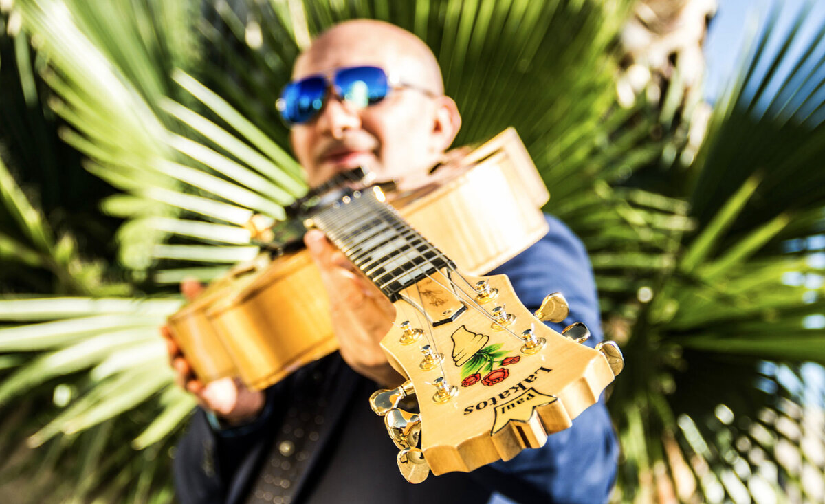 Male musician portrait Paul Lakatos wearing sunglasses pointing yellow wood guitar towards ground palm tree backdrop