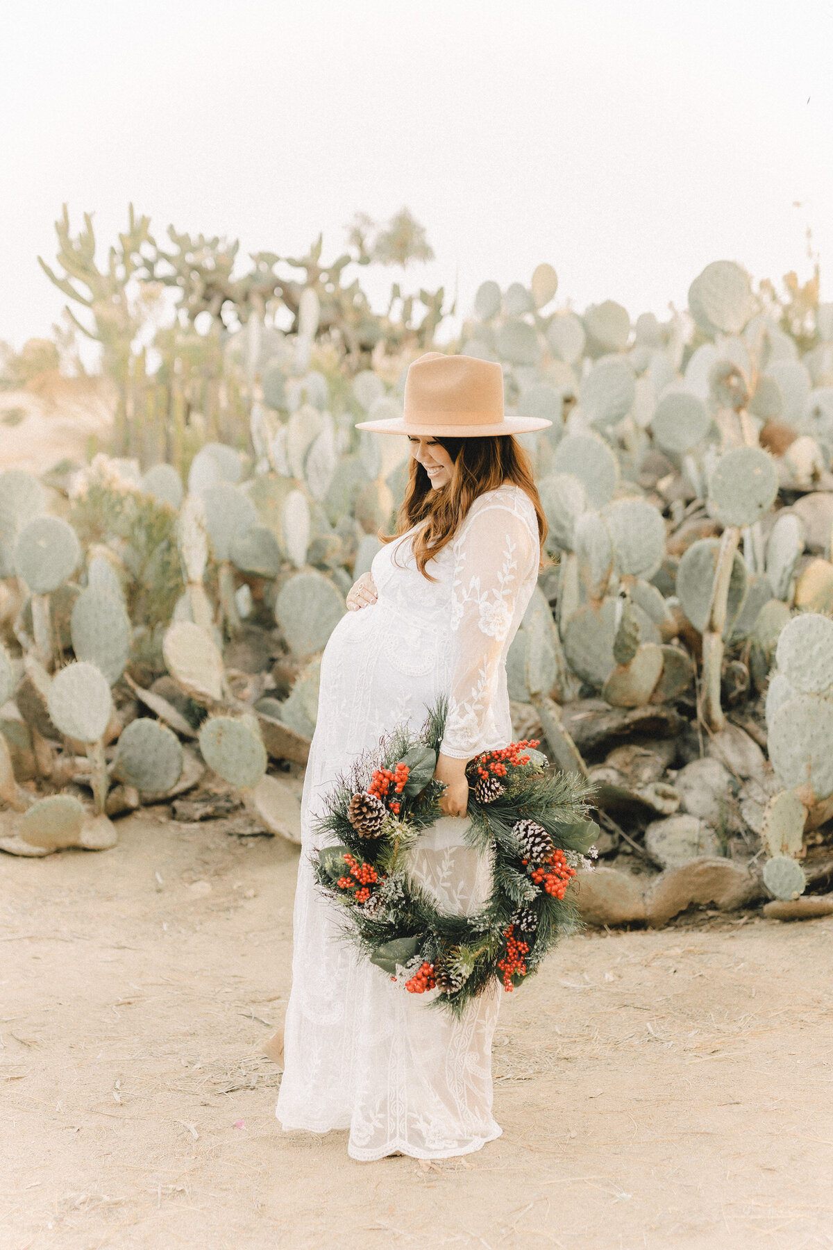 Pregnant woman in a white lace maternity dress and tan hat holding a festive holiday wreath, standing in front of desert cactus plants in San Diego.