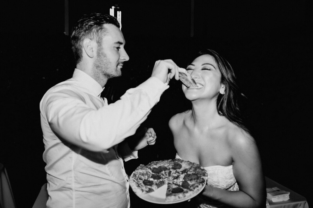 man in collared shirt and bow tie feeding a woman in a wedding dress pizza, she is very excited