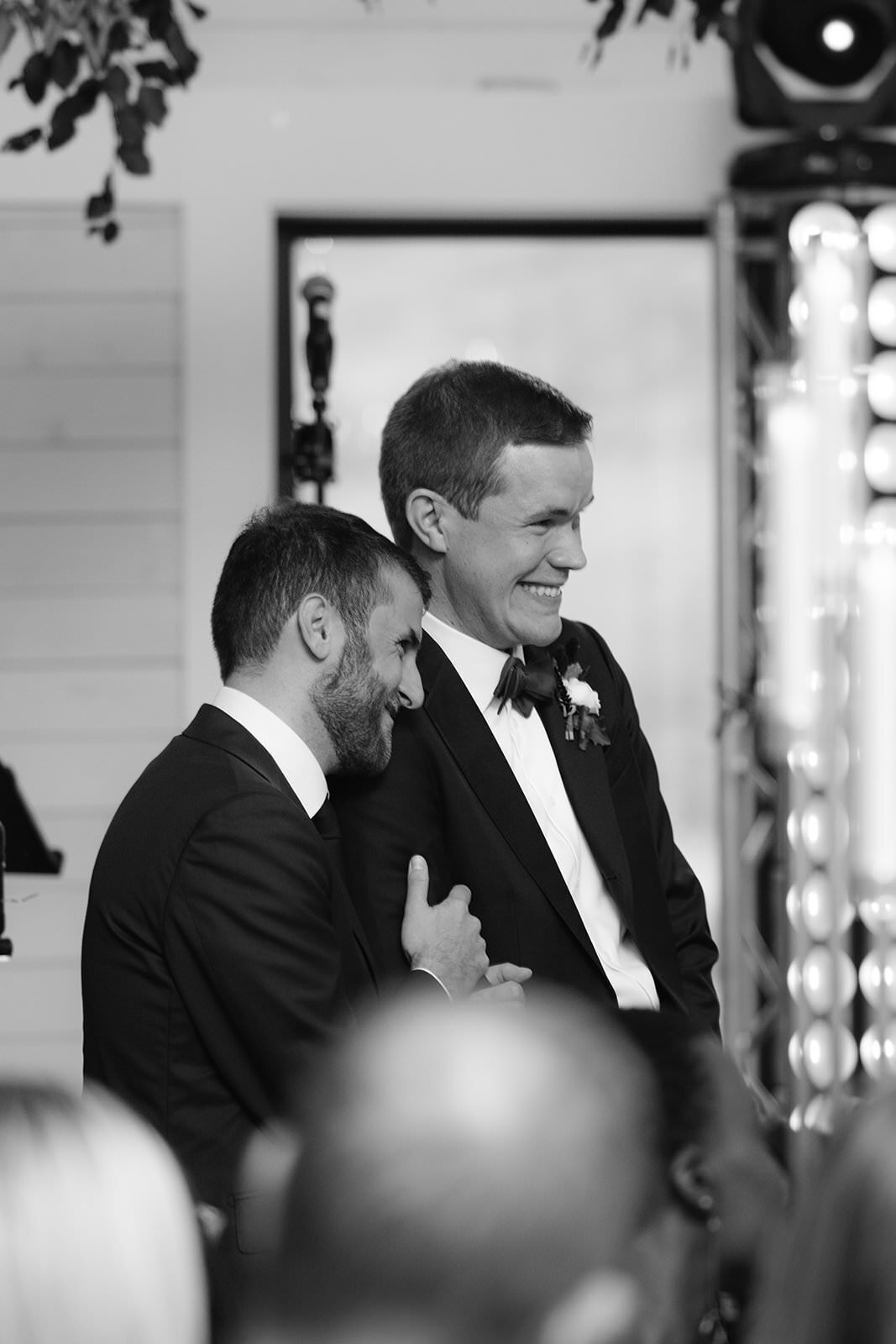 Two grooms hold each other smiling during wedding toasts