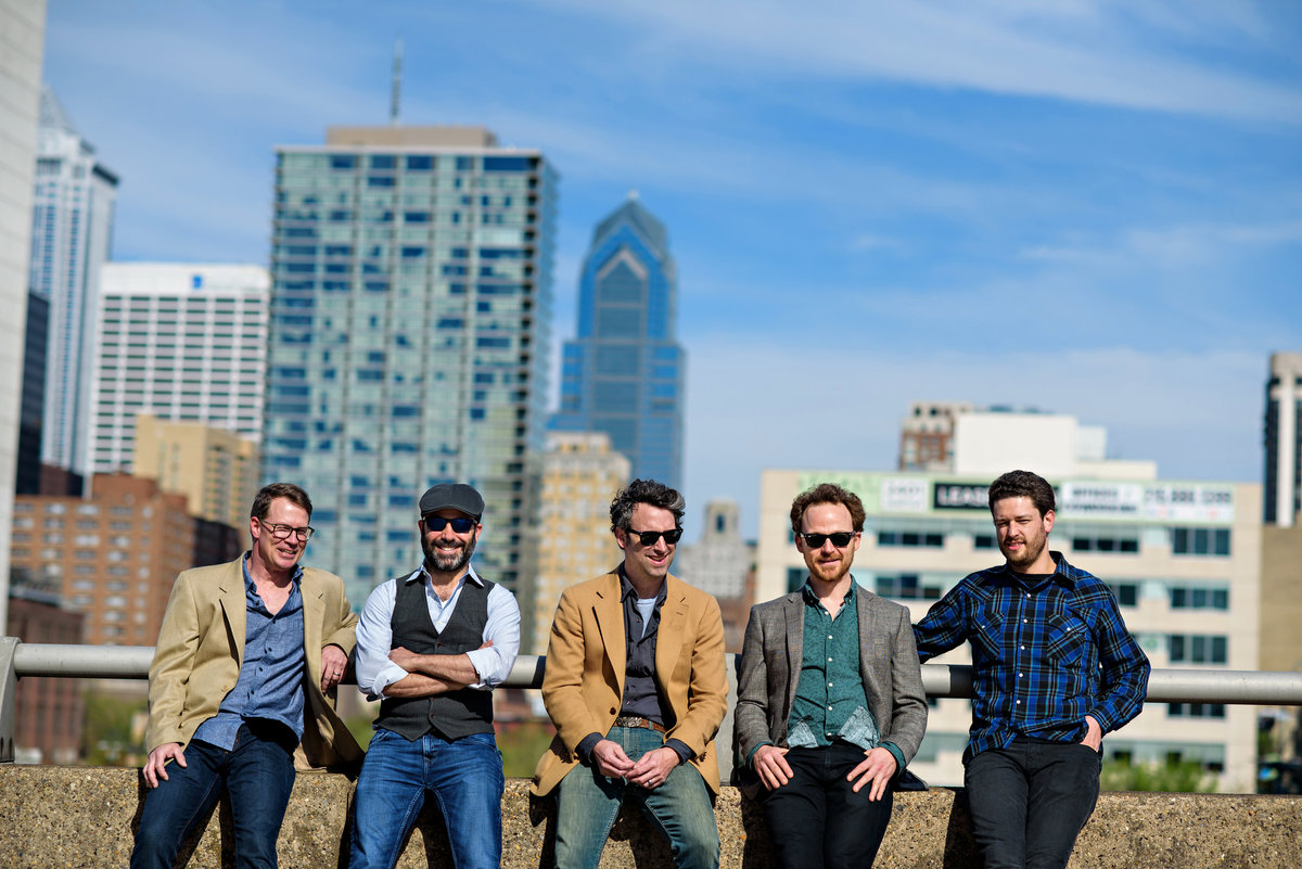 A Philadelphia band called Marc Silver and the Stonethrowers sit on a wall with the philly skyline behind them.