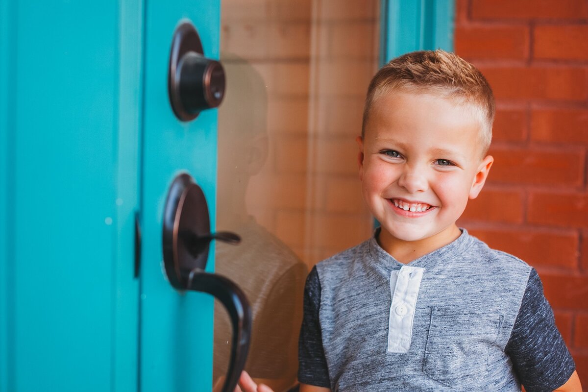 young-boy-with-short-blonde-hair-smiling-by-a-vibrant-blue-door-in-downtown-fortville-indiana-during-a-lifestyle-family-photography-session