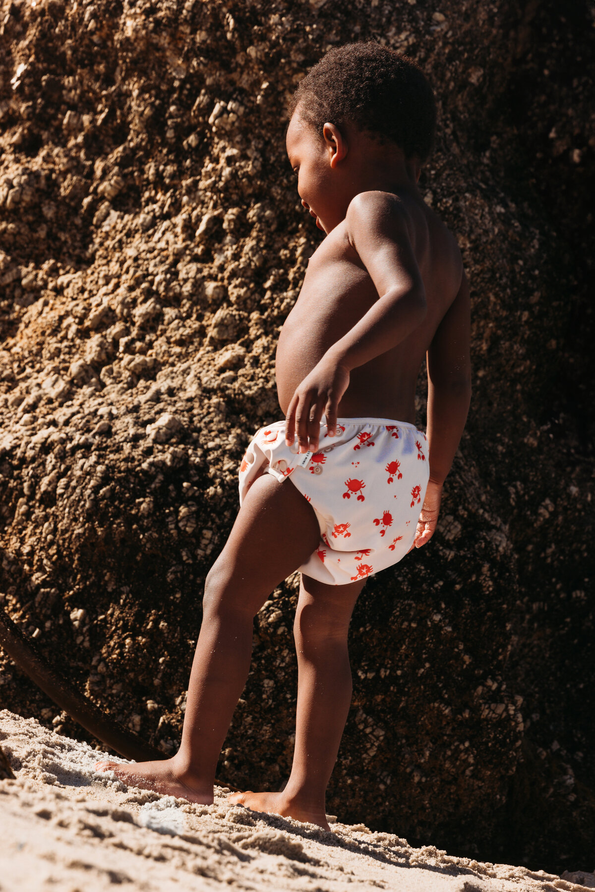 Young child in a Swim Essentials swim diaper with red crab print, standing barefoot on the beach near a sunlit rock formation in Cape Town.
