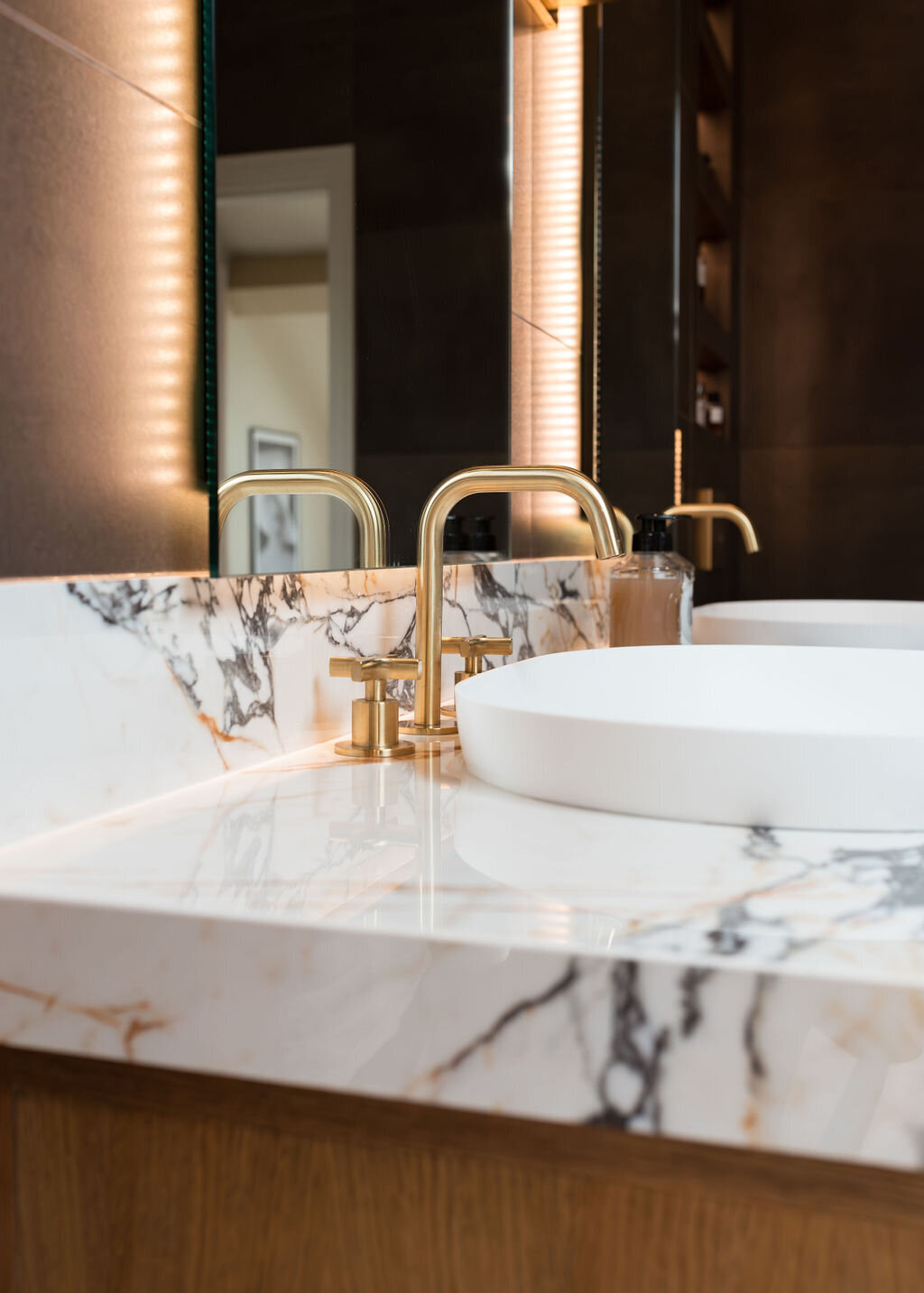 A modern bathroom, featuring a double sink vanity with marble countertops and wooden cabinets.The room is finished with dark tiled walls and light tiled floors.