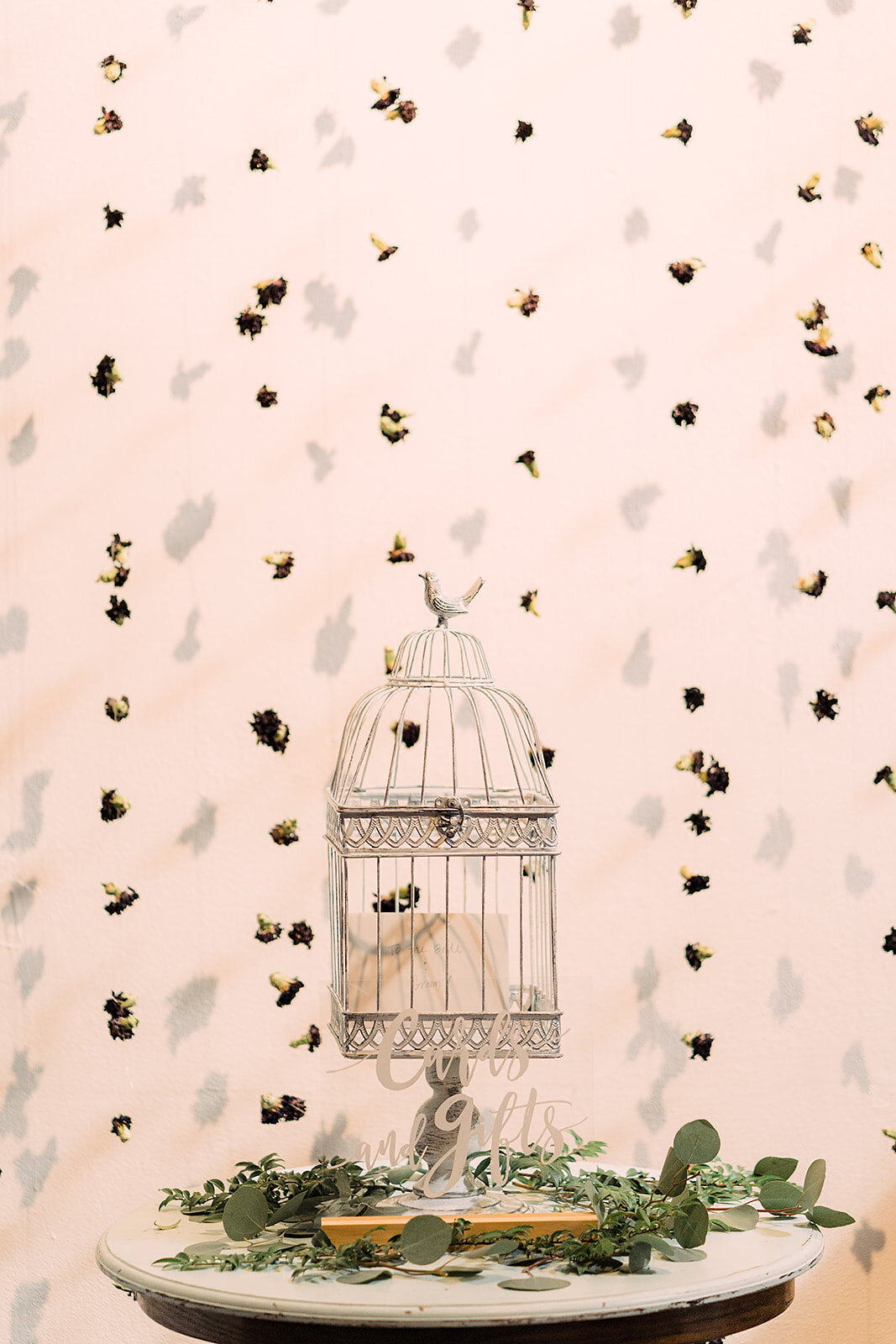 A birdcage cardholder labeled "Cards and Gifts" sits on a round table, adorned with greenery and surrounded by a backdrop of small dried flowers scattered across a light pink background.