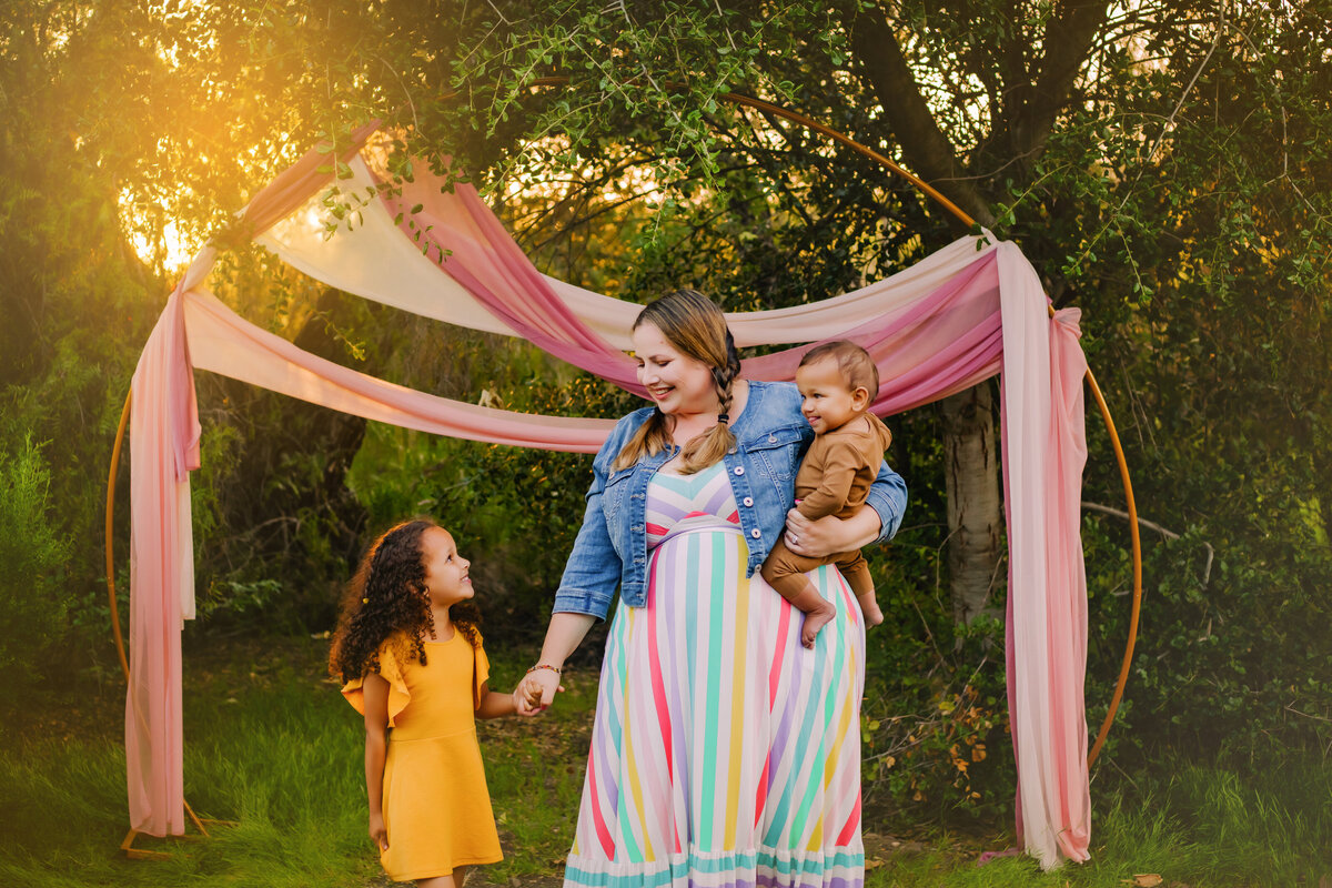 Family Photographer,, mom walks with her daughter and baby outside beneath colorful drapes