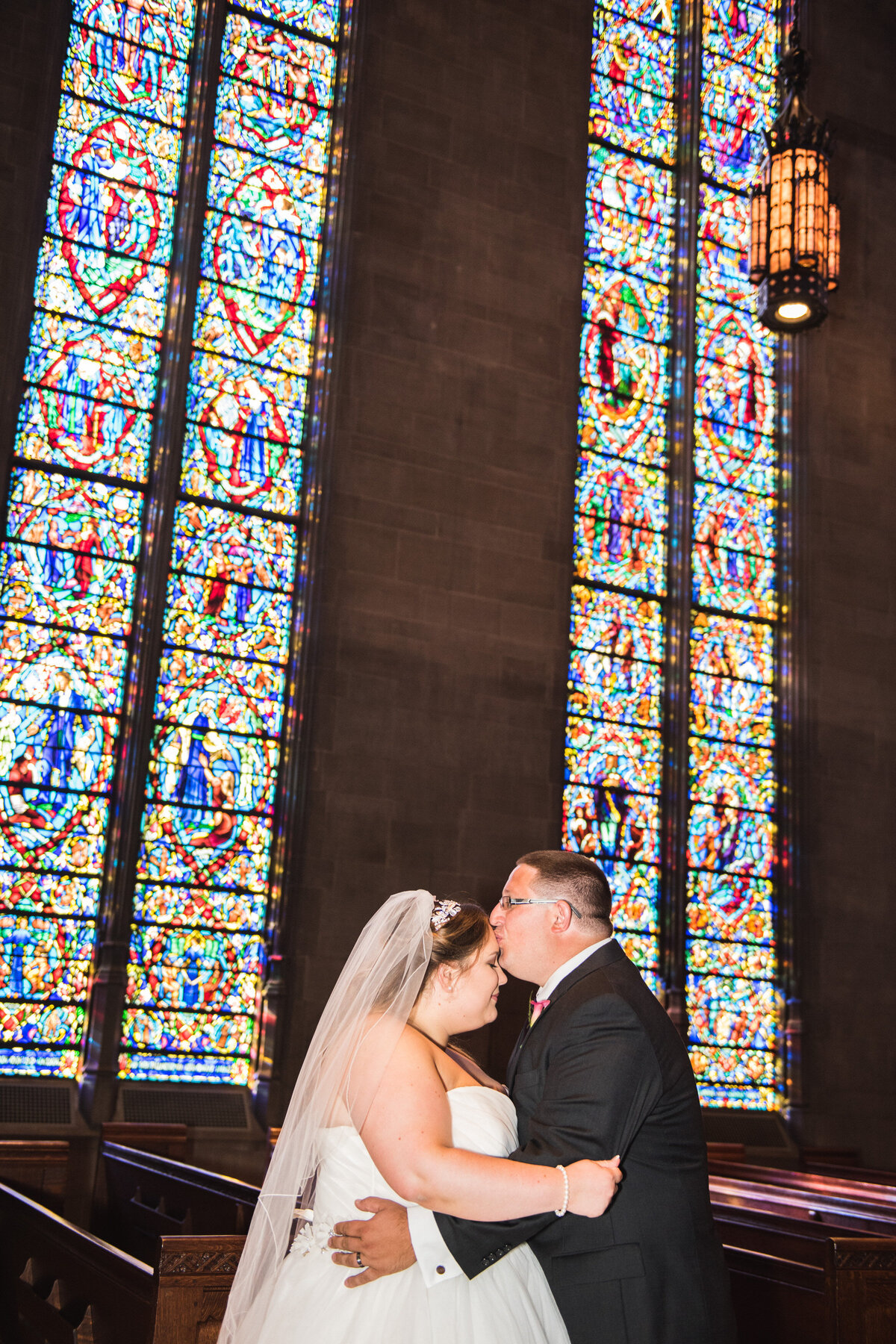 a person kissing their partner's forehead with stained glass windows behind them 