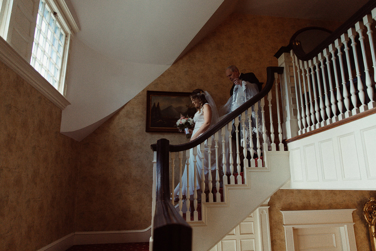 bride walks down stairway with her father holding her dress at Dibbles Inn