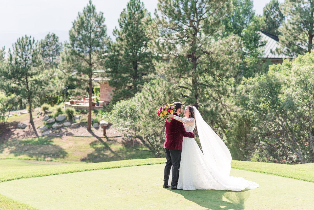 Bride and groom stand on a golf course with her arms and flowers over his neck while he brings her in close to him and then face each other captured by Denver wedding photographer