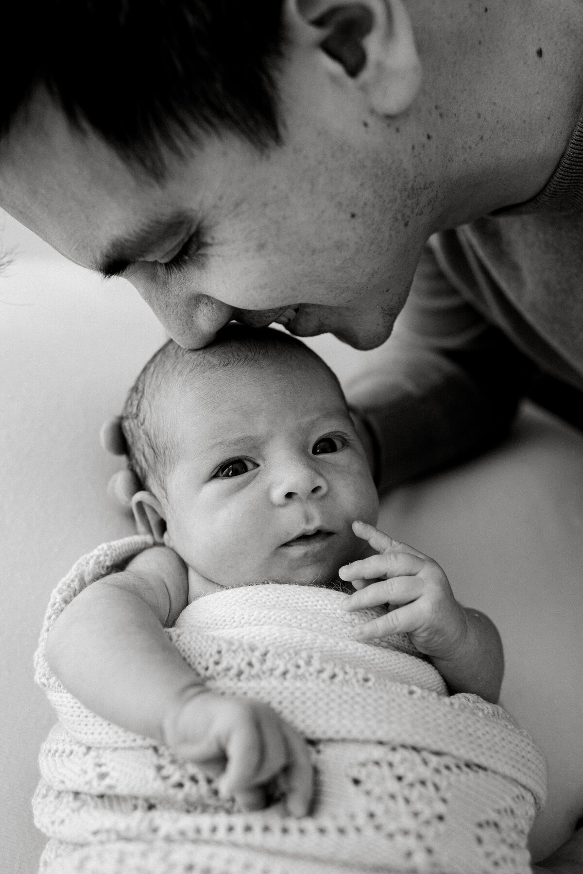A father kisses his newborn baby while the baby looks at the camera and is photographed by lucy jane photography in her christchurch newborn photography studio
