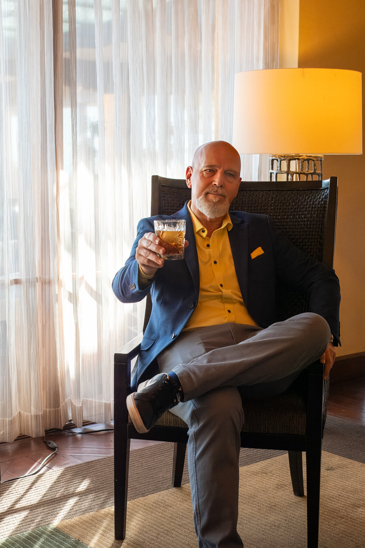 Man sitting confidently in a chair holding a drink, wearing a blazer and yellow shirt in a warm indoor setting, photographed by Vyrl Photo for Tucson brand photography.