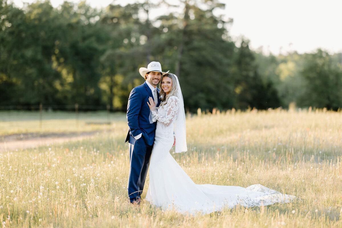 Groom in navy suit and straw cowboy hat standing in a field of wildflowers with bride in traditional lace wedding gown and long pearl beaded veil after East TX wedding ceremony