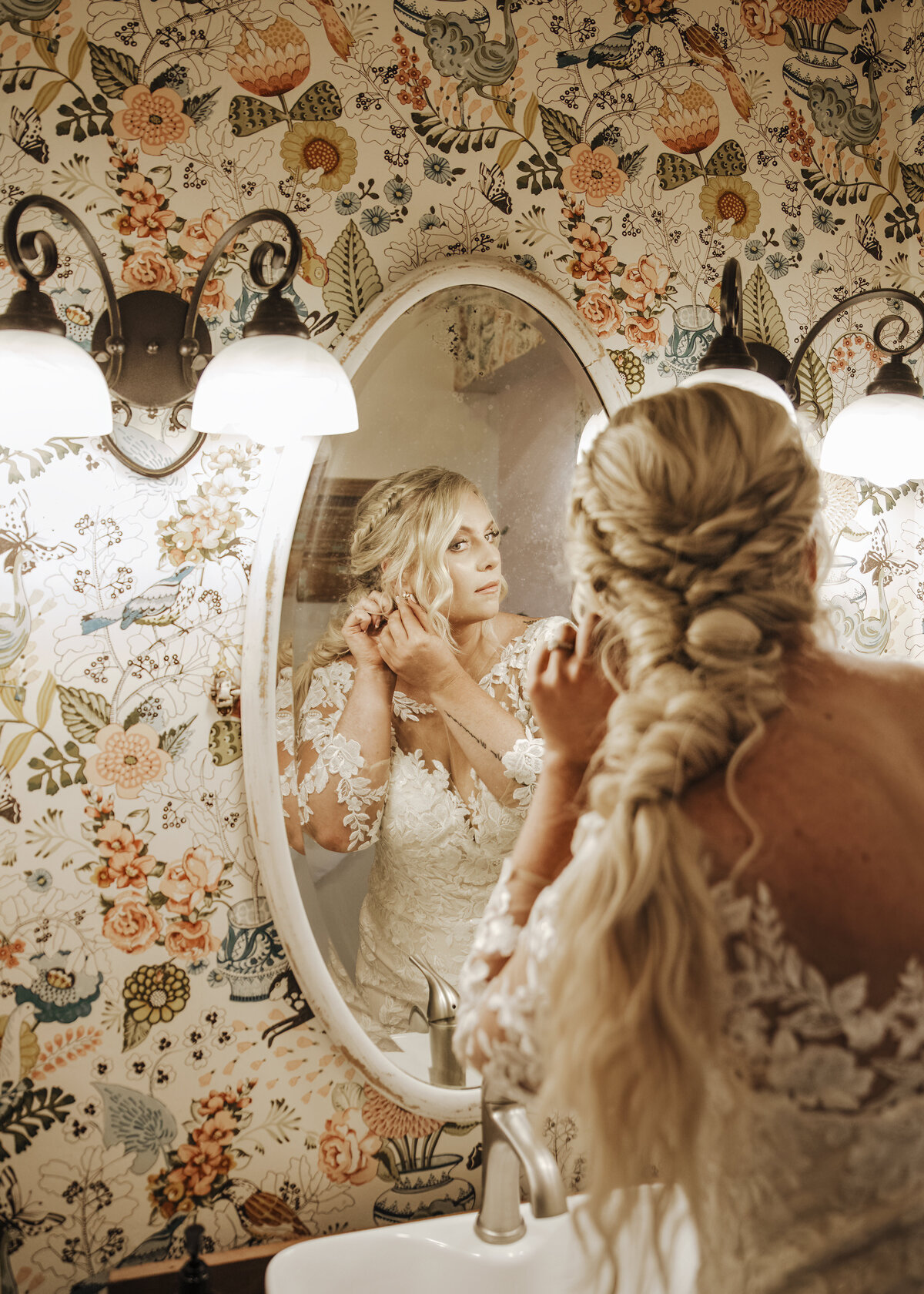 A bride in a lace gown finalizing her look in front of a mirror with ornate floral wallpaper in the background taken by jen Jarmuzek photography a Minneapolis wedding photographer