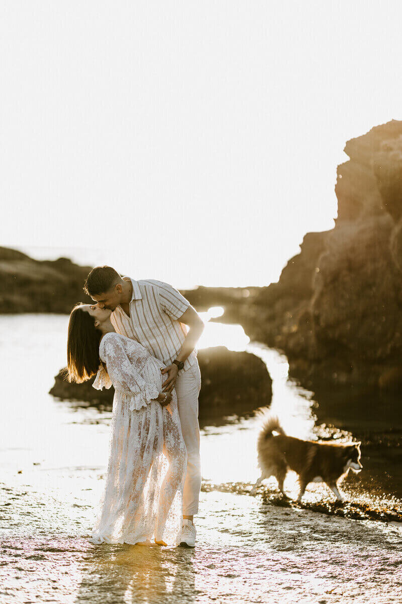 Couple s'embrassant entouré de leur chien et de rochers, au coucher de soleil, pieds dans l'eau. L'homme caresse le ventre rond de sa femme enceinte. Photo prise par Laura, photographe professionnelle.