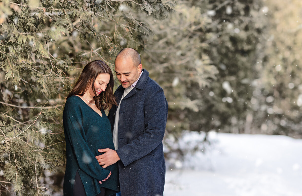 A beautiful outdoor winter maternity portrait photographed after a major snowstorm at Centennial Park in Ayr, Ontario. The expectant couple is surrounded by evergreens as snow gently falls.