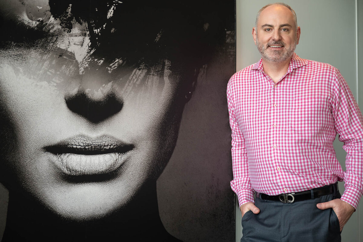 A portrait of a man standing next to an wall art of a woman's face in black and white. The man is wearing a red and white plaid shirt and blue pants