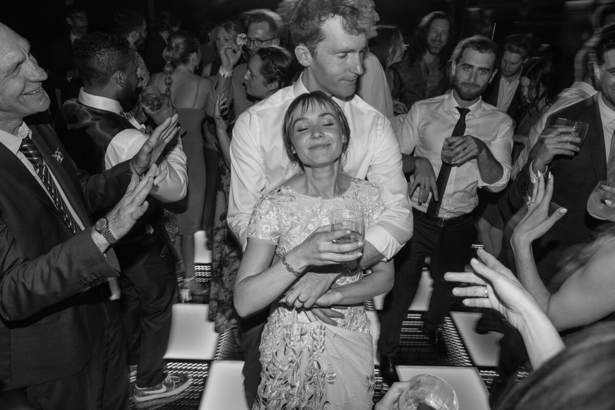 Groom embraces his bride while they party with their friends and family in black and white